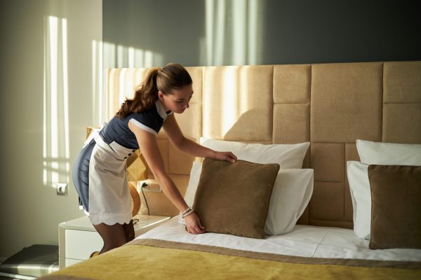 Housekeeper arranging pillows on a neatly made bed in a hotel room with sunlight streaming through window blinds